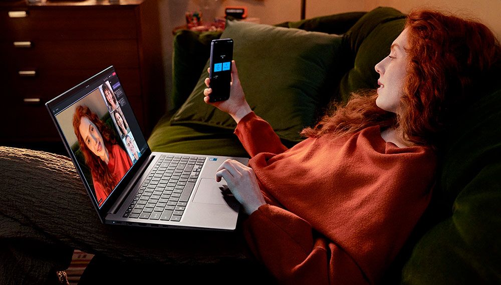 Mujer usando un smartphone junto a un portátil Samsung Galaxy Book4 en una estancia con luz tenue.