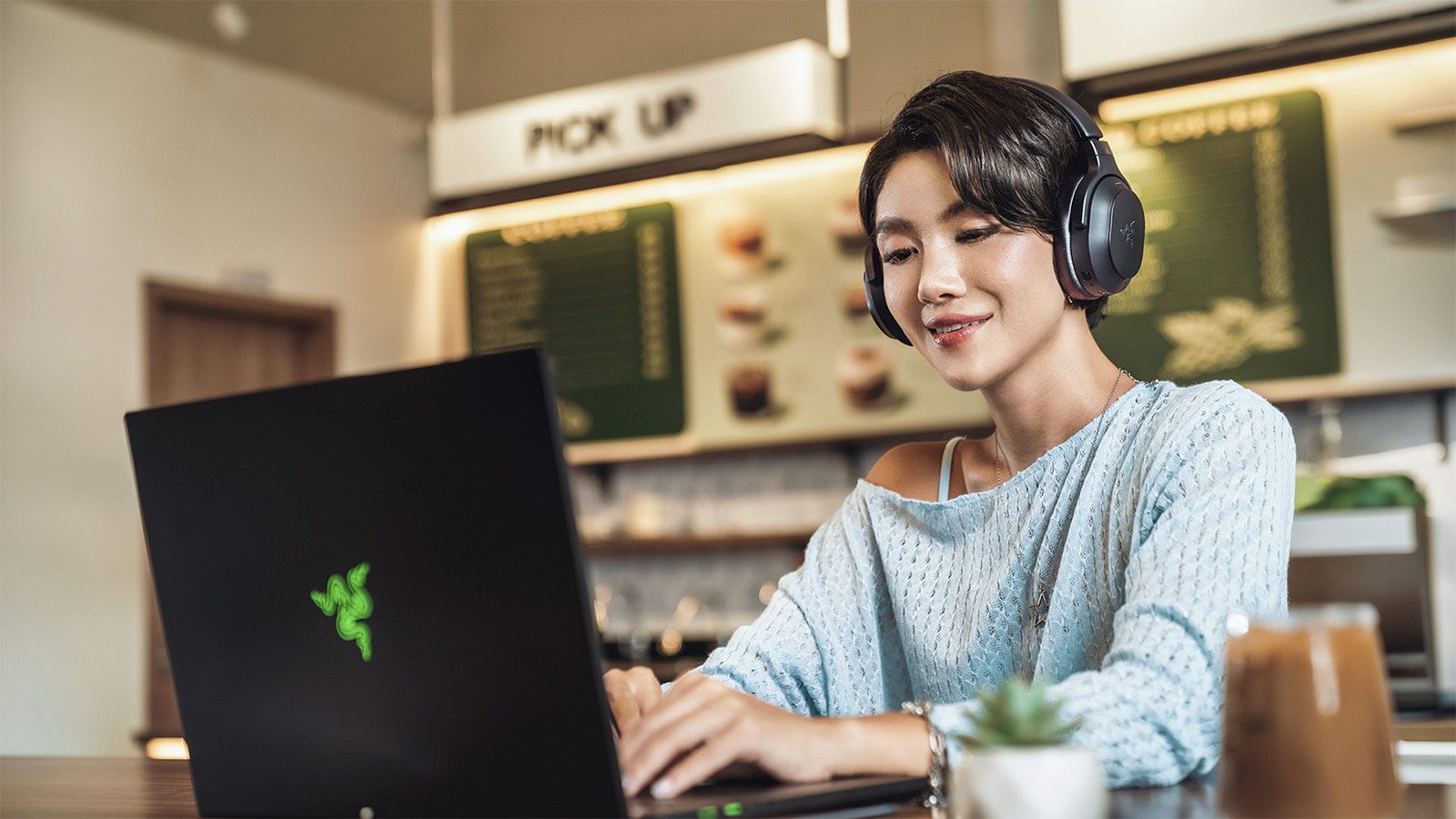 Mujer usando auriculares Razer Barracuda v2 Pro mientras trabaja con un portátil en una cafetería.