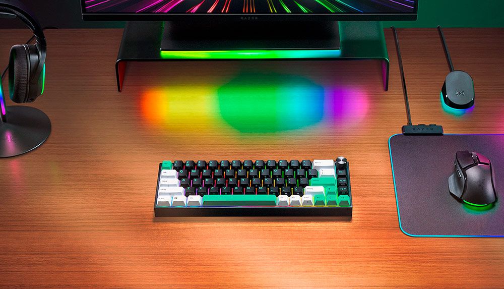 Teclado Razer Reclusa Mini en un escritorio de madera con iluminación RGB proyectada desde el monitor y periféricos de juego.